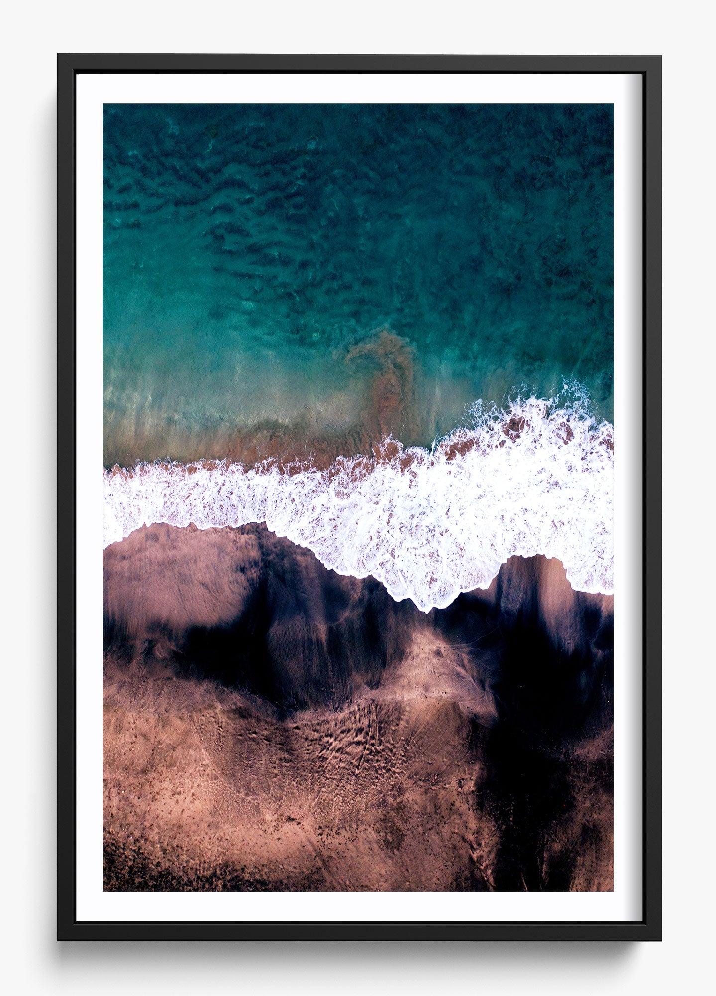Framed wall decor featuring an aerial view of a beach with dark sand, turquoise water, and white foamy waves.